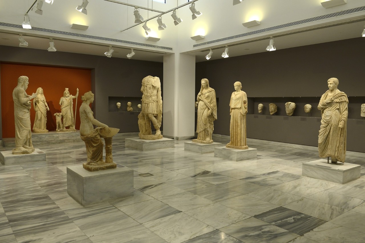 The picture shows eight human sculptures from ancient Greece, each standing on a pedestal. They are in a room tiled with marble. The wall painting is partly grey and partly red. In the grey areas of the wall, niches contain busts.