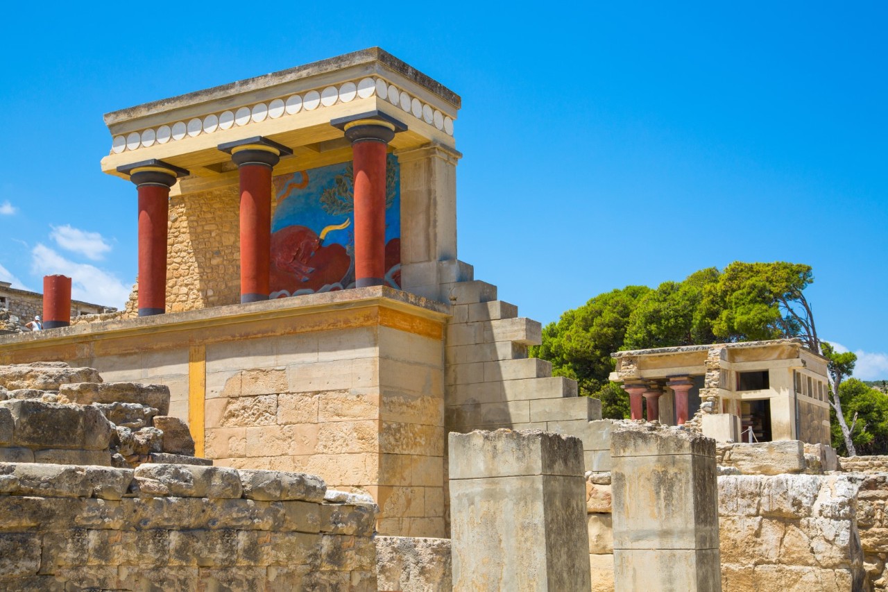 View onto the ruins of a beige-sand-coloured building with three red columns on a pedestal. These support a flat roof. There are trees in the background. The sky is a bright blue. 
