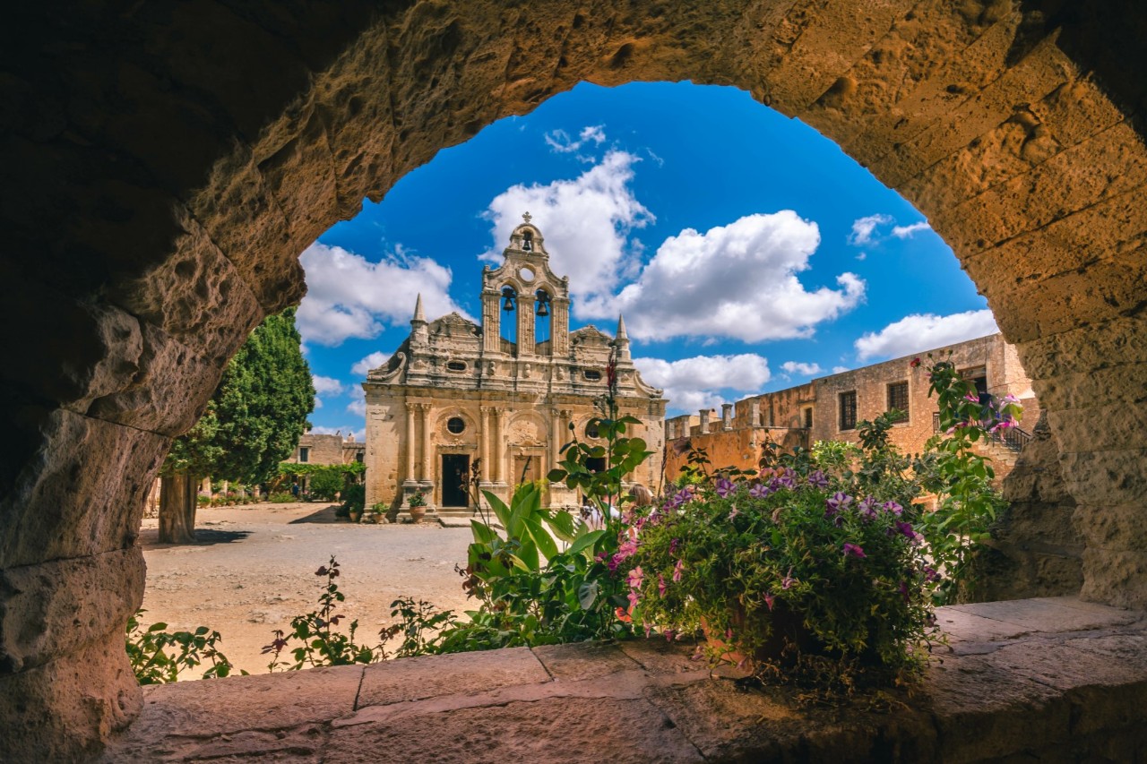 View through an old stone arch window onto a historic sand-coloured monastery. Green plants grow below the window. The view leads to an imposing portal decorated with curves, arches and towers. To the left of the portal stands a tree, to the right are further buildings. The sky is a bright blue with white clouds.