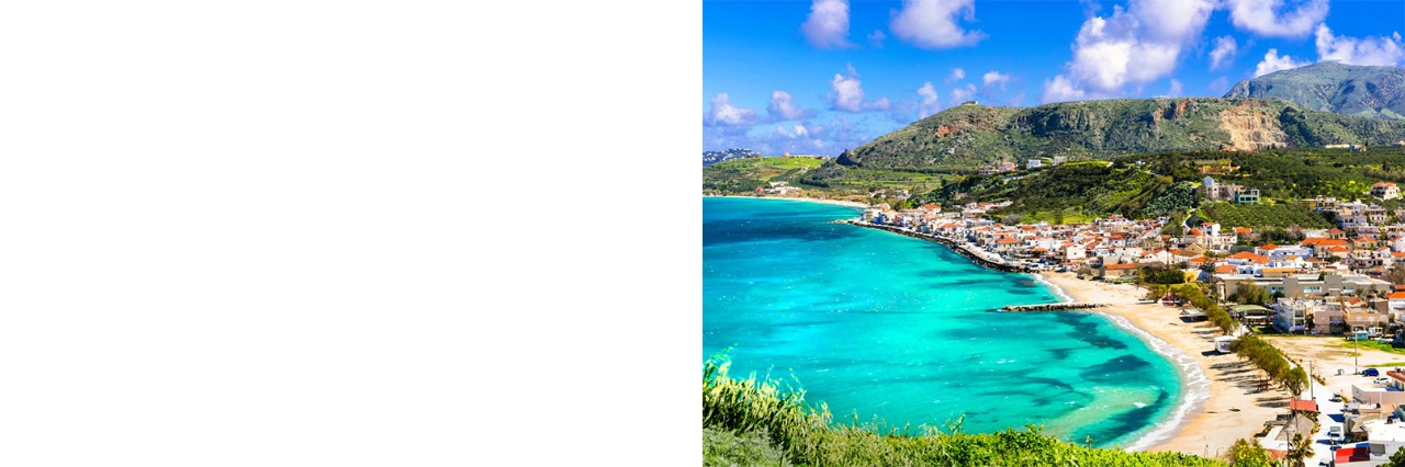 A green mountain over a wide bay with a sandy beach on the right. Behind it is a town with many white houses and red roofs. The hinterland is hilly to mountainous with green vegetation. The water is turquoise with a few dark blue highlights. The sky is blue with white clouds. 