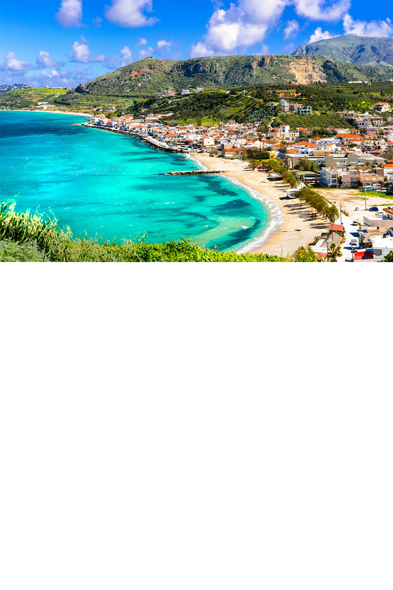 A green mountain over a wide bay with a sandy beach on the right. Behind it is a town with many white houses and red roofs. The hinterland is hilly to mountainous with green vegetation. The water is turquoise with a few dark blue highlights. The sky is blue with white clouds. 