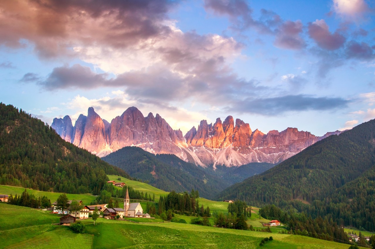 A reddish glowing mountain range in the centre, with further green mountains in front of it. In the foreground, there is a large, green alpine meadow and a small mountain village with a church.