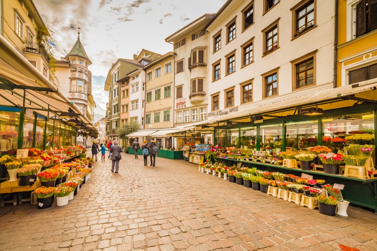 Cobbled old town lane with old, multi-storey houses on both sides. Both sides of the lane are lined with market stalls selling fruit, vegetables, and flowers. A few people stroll in the lane.