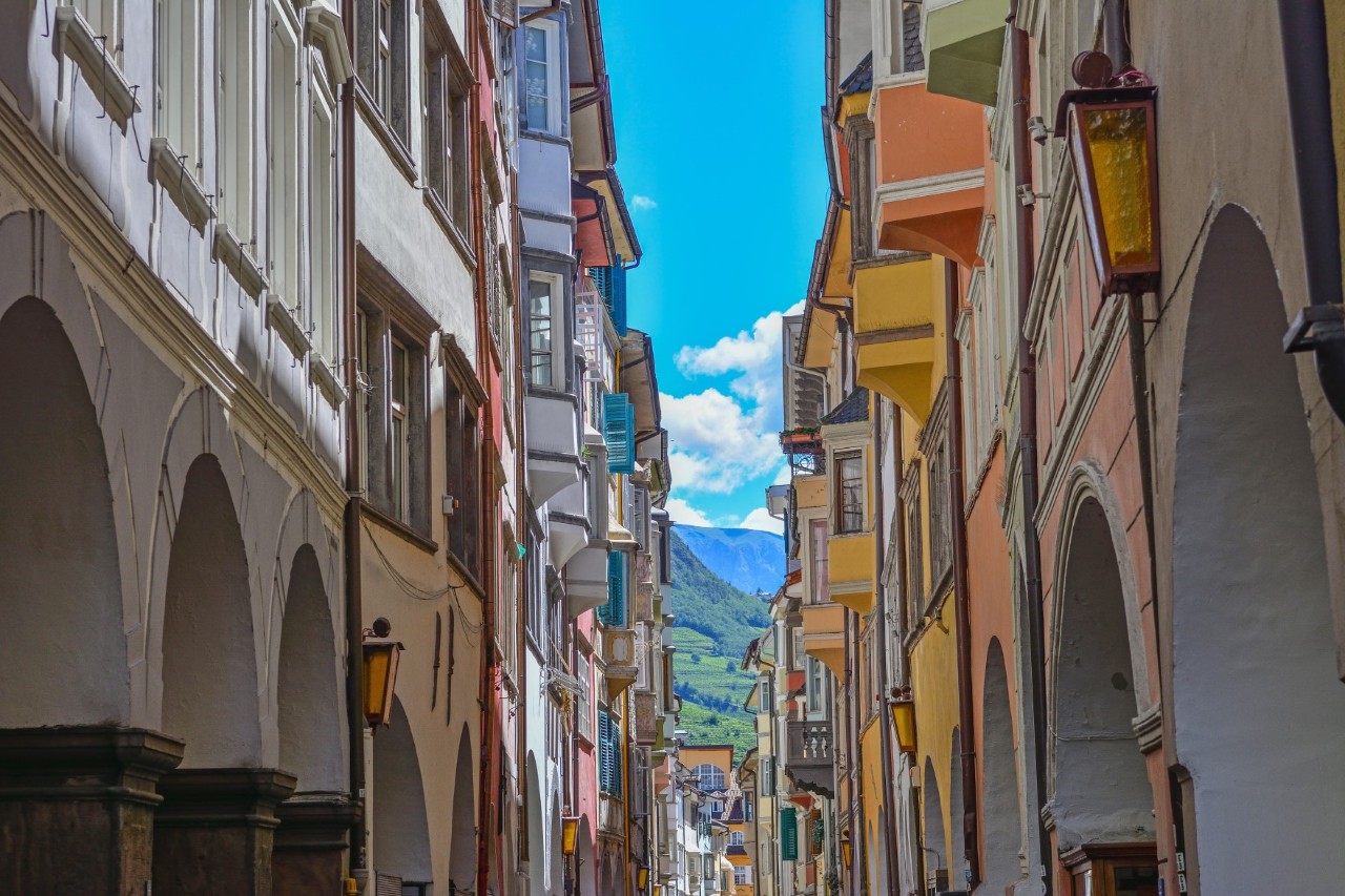 View of a narrow alley with colourful, multi-storey houses on both sides with small oriels and open archways. A mountain is visible in the background.