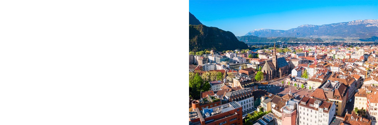 View of the city from above, numerous houses with red tiled roofs can be seen. In the centre stands a striking church. Mountains can be seen on the left and in the background.