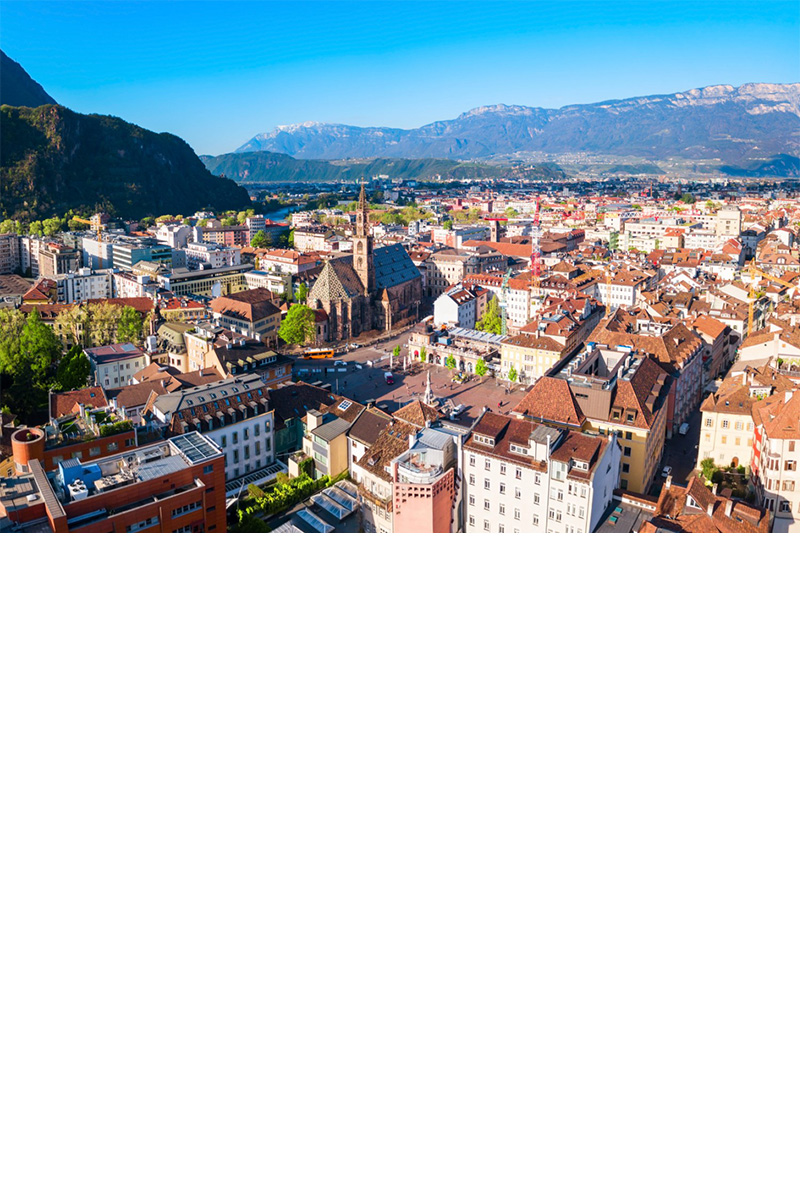 View of the city from above, numerous houses with red tiled roofs can be seen. In the centre stands a striking church. Mountains can be seen on the left and in the background.