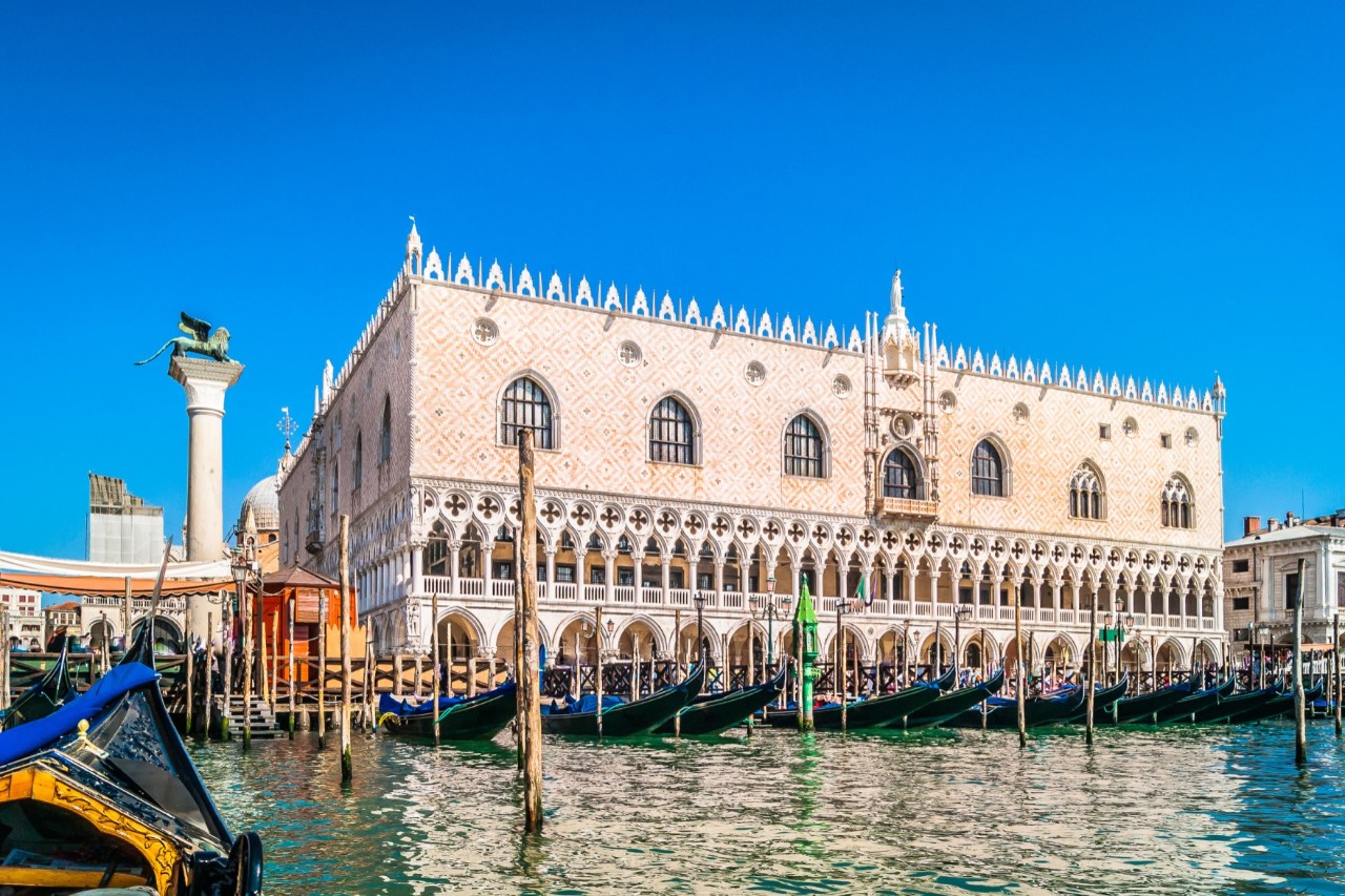 Doge's Palace on the Grand Canal, in front of it canal with a small harbor, Venetian gondolas, masts in the water, blue sky. © dreamer4787/stock.adobe.com