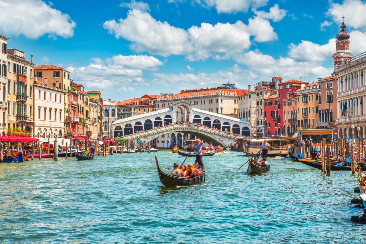 Grand Canal, palazzi on the left and right, gondolas with tourists and gondoliers, Rialto Bridge in the background, sunshine. © Yasonya/stock.adobe.com