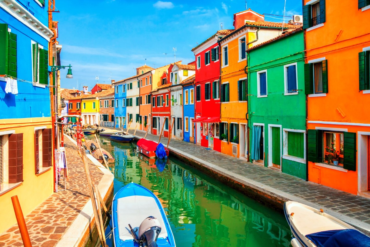 Colourful houses line the narrow canal, whose water has a greenish tinge. Small boats are moored on the deserted bank. The sky is a bright blue.