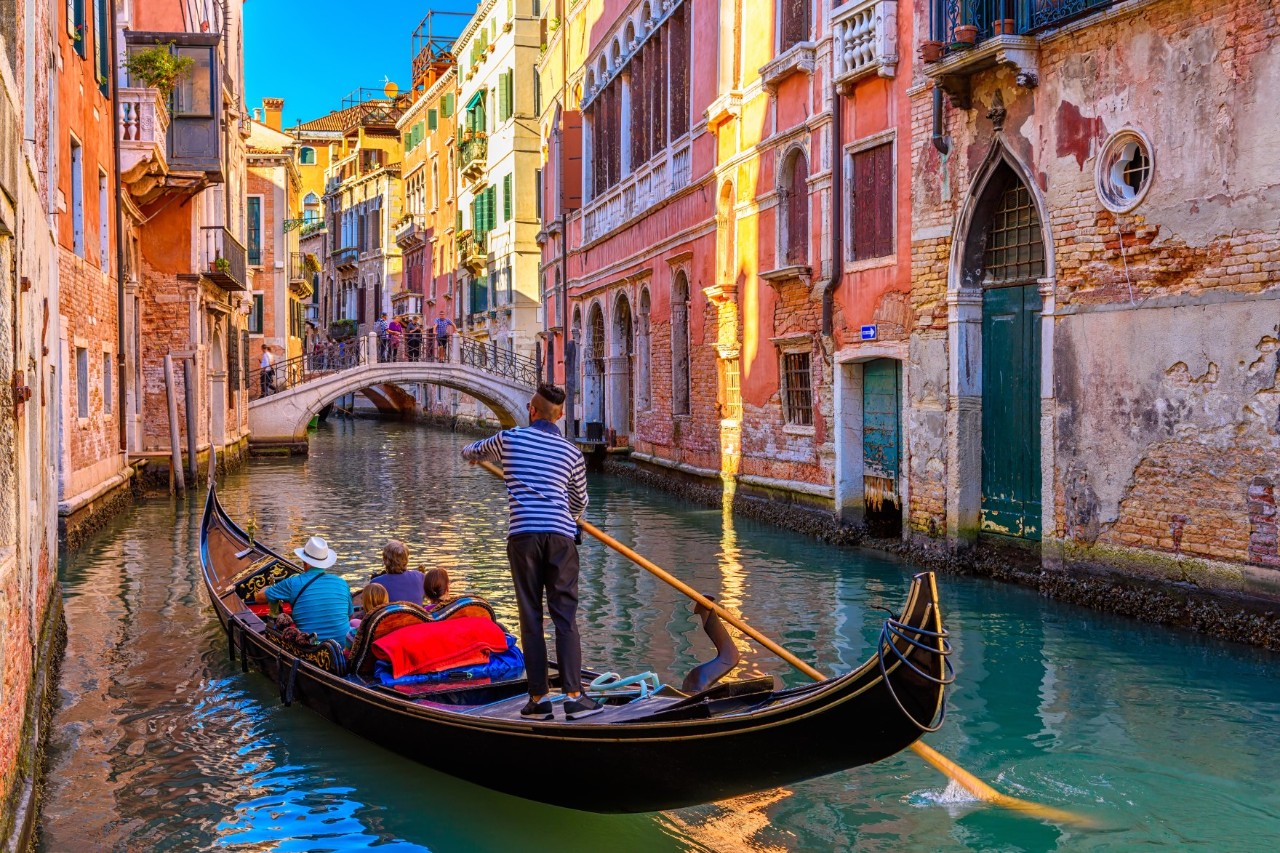 A gondola passes through a narrow canal with turquoise blue water. The house facades glow in yellow and orange and in the background there is a small bridge.