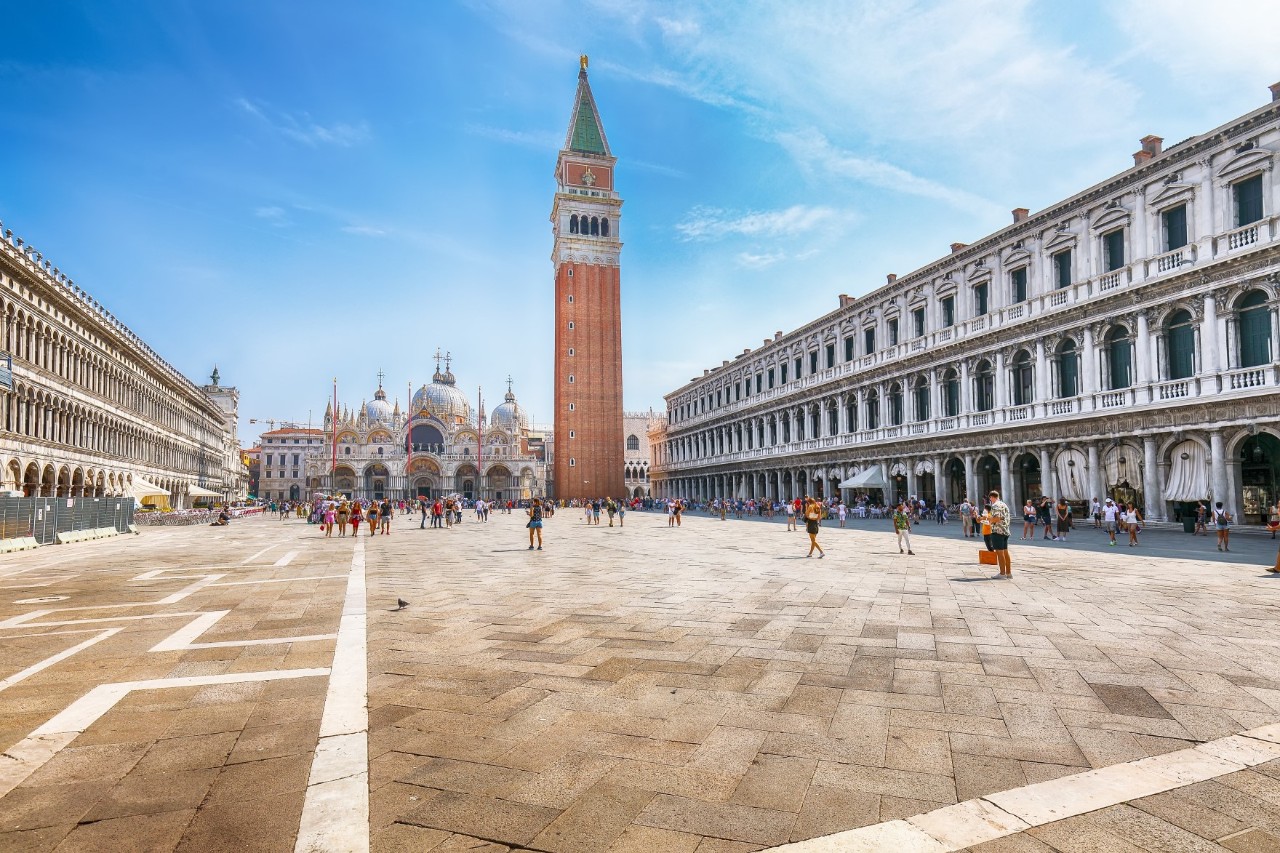 A large, reddish-brown tower with a green spire and a basilica with numerous domes behind it. In the foreground, there is a large square with few people. To the left and right, there are elongated buildings with lavishly decorated façades.