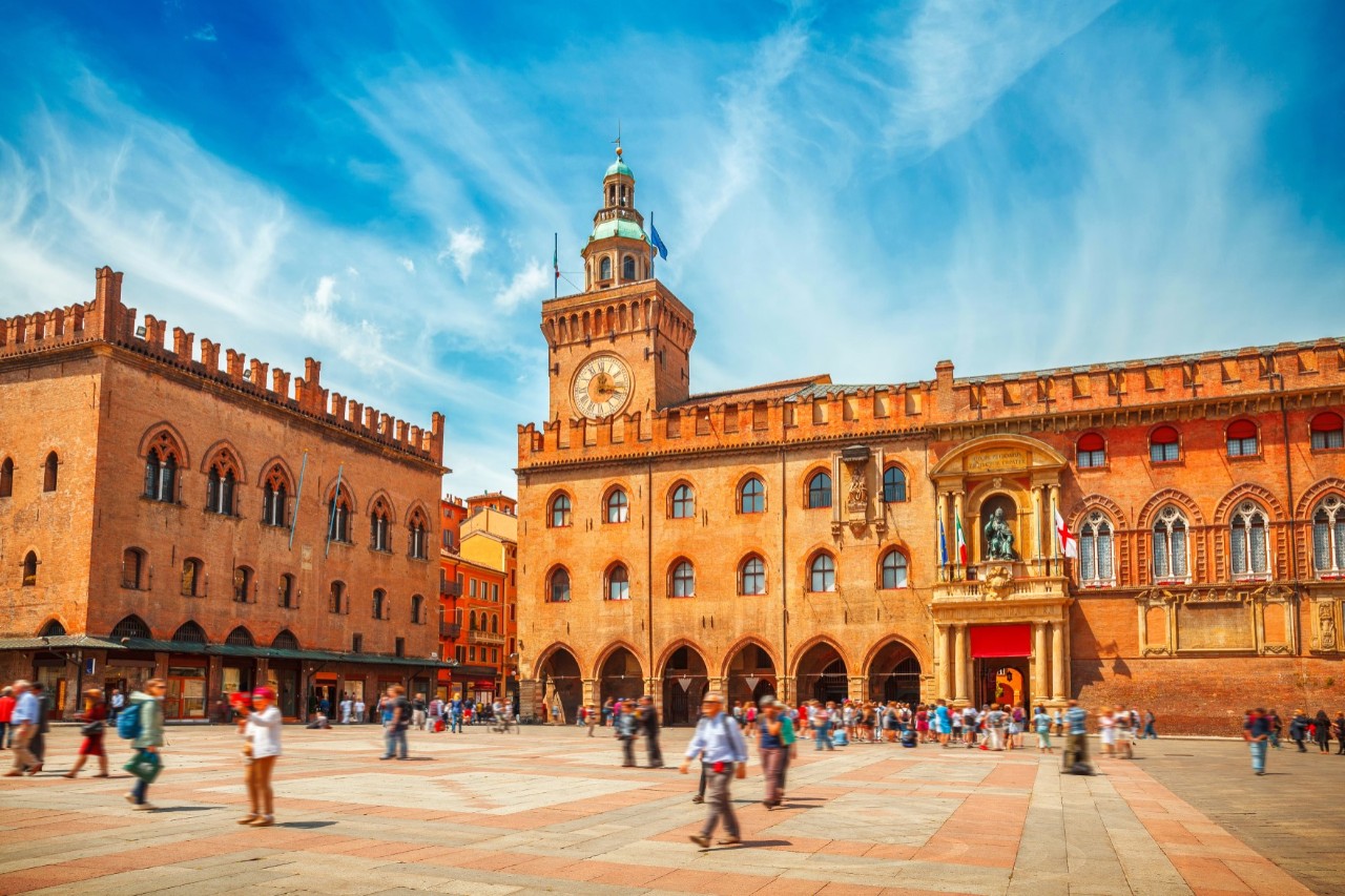 A large square, the floor of which is paved with large red and grey tiles. Numerous people can be seen on it. Behind them are two large, brick-coloured buildings, both with crenellated roofs. A small, square clock tower rises from the right-hand building to the left. An alley leads between the buildings. The sky is slightly cloudy and blue.