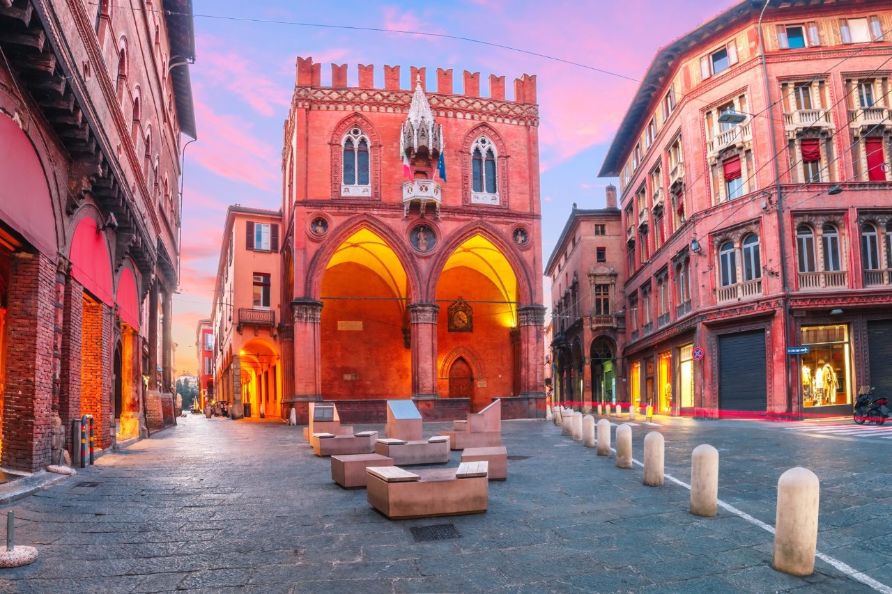 In the red evening sky rises a narrow, red, historic building with two high, pointed arched entrances. The roof is crenellated. There is a traffic-calmed square around the building, separated from traffic by bollards. A large sculpture stands in front of the building. On both sides of the building, other historic buildings can be seen shining in the evening light.