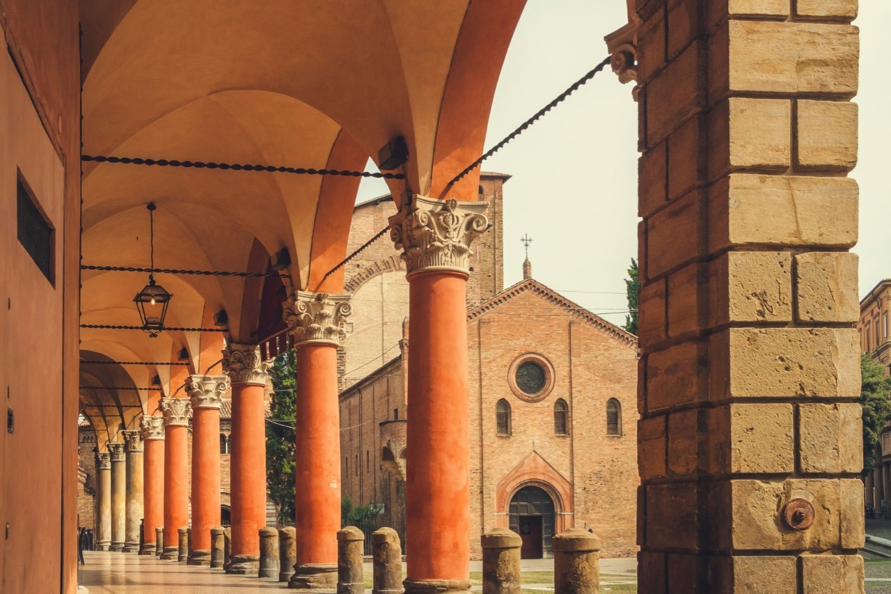 An archway with red columns with decorated capitals. There are bollards between the columns. A church with a pointed nave can be seen behind the columns.