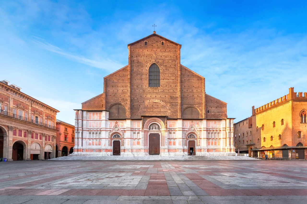 Frontal view of a large, massive cathedral standing on a red and grey tiled square. The lower part of the façade is decorated in pink and white, with three doors forming the entrance to the cathedral. The upper half of the façade is made of rough brick. The nave is rectangular, the gable is triangular and tapers upwards. There are historical buildings on both sides of the cathedral, the one on the right is illuminated by the sun.