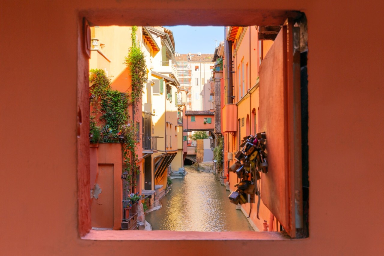 View through a small window set into a red façade. The view opens onto a narrow canal with colourful house facades on either side. A small covered bridge over the canal is visible in the background, with more houses behind it.