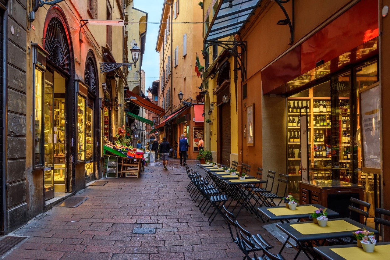 View of a narrow alley in an old town centre in the evening light. On both sides are multi-storey houses with shop windows illuminated at ground level. On the right-hand side of the alley are the tables of a restaurant, with wine bottles on shelves in the window behind them. A vegetable stall and other shops with awnings can be seen at the back of the alley.