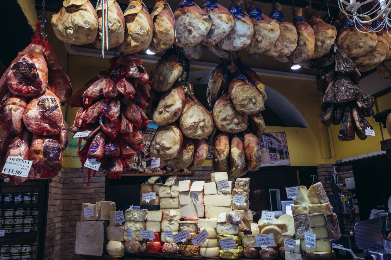 A market stall full of culinary treasures: Numerous hams of different varieties and sizes hang from the ceiling, while cheese wheels and pieces of cheese are piled high in the display.