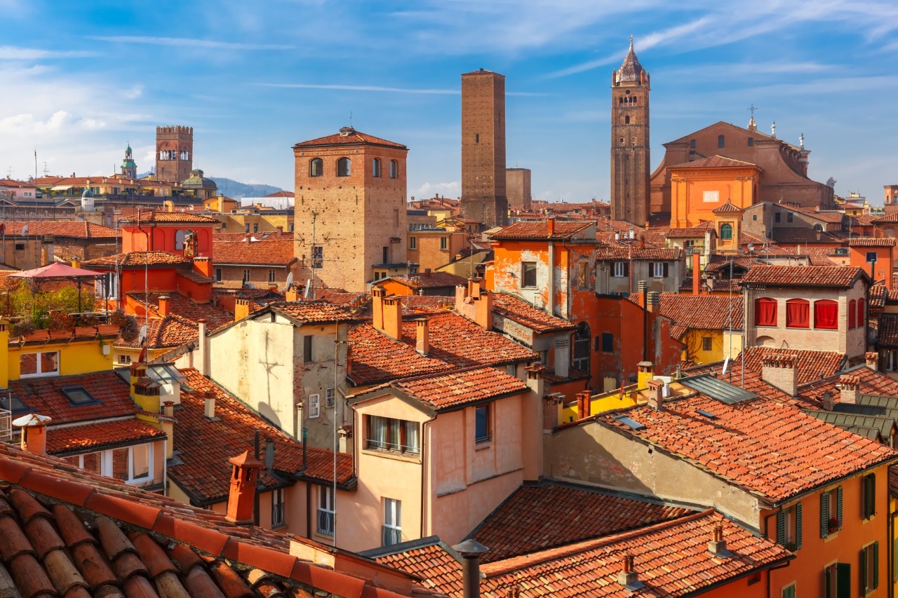 Aerial view of an old town centre in which red-tiled roofs and reddish facades dominate the scene. Several square towers rise between the houses. The roofs glow in the sun.