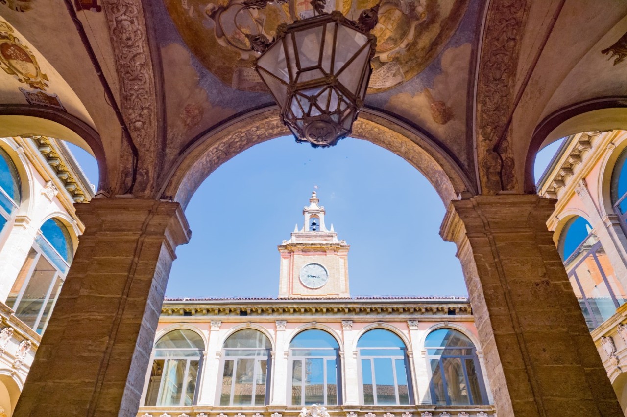View upwards from an arcade onto a surrounding building with high arched windows, on which a pink-coloured clock tower stands in the centre. The arcade from which the picture is taken is decorated with a ceiling painting and a historic lantern. The columns on either side are angular and made of brick. The sky is blue.