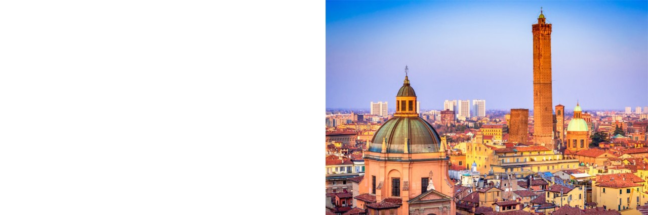 Aerial view of a large city in the reddish evening light. In the foreground is a large, round church dome behind which are numerous houses with tiled roofs and historic buildings as well as two adjacent square towers, one of which is significantly higher than the other. 