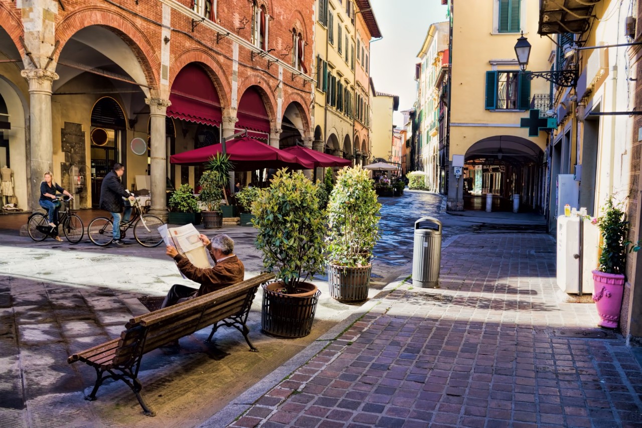View of a small square in the centre of Pisa, lined with magnificent old buildings with arcades. A man reading a newspaper sits on a bench in the foreground, while cyclists and the parasols of a café can be seen in the background 