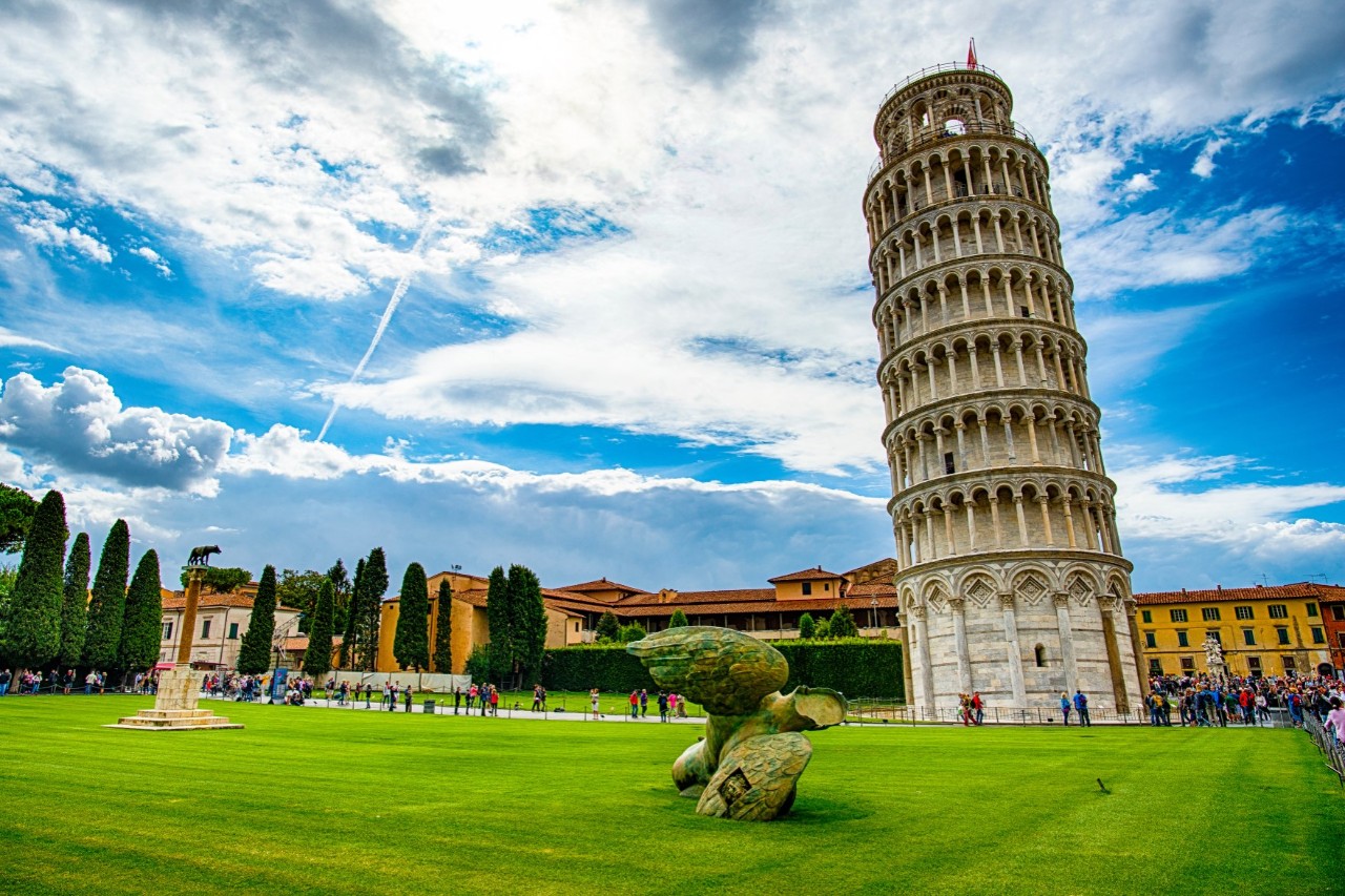 View of a leaning tower from a extensive lawn with a sculpture and a monument. There is a gallery of columns surrounding each floor of the round, high tower 