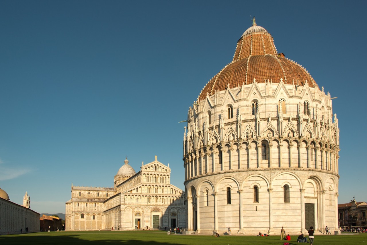 Frontal view of a large, round baptistery made of white marble. People can be seen on the lawn in the foreground and Pisa Cathedral in the background 