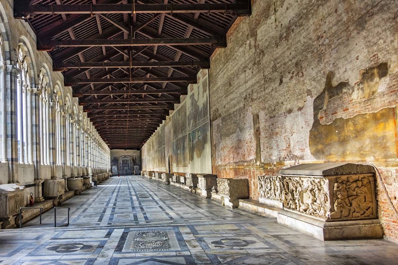 An arcade. The walkway is lined with stone sarcophagi, the open arcade arches can be seen on the left and fragments of frescoes on the right-hand wall 