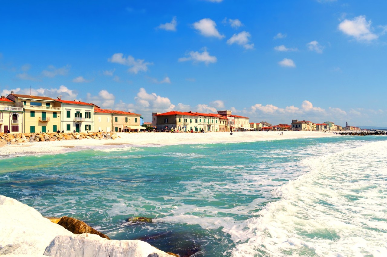 Gentle waves break onto a white sandy beach that runs out to the right into a pier, behind the beach are brightly coloured buildings with red roofs 