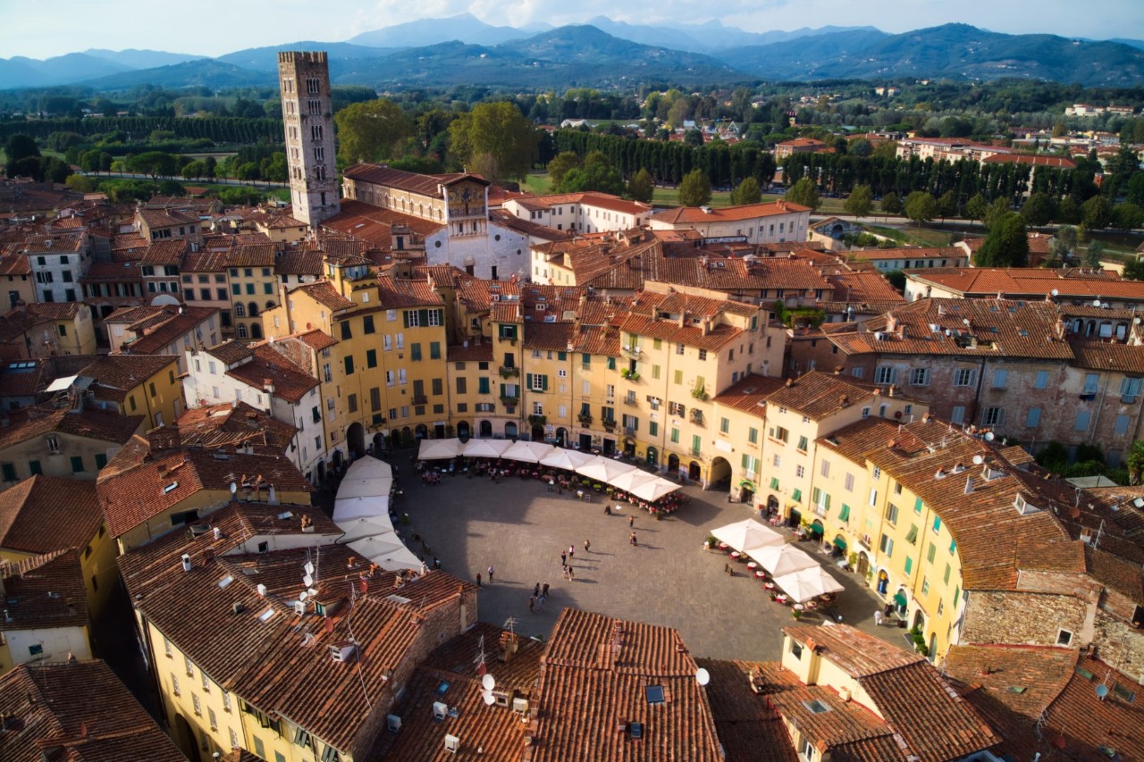 View from above of an oval square lined with old buildings where market stalls can be seen. There is a tower at the far end and a hilly landscape on the horizon 
