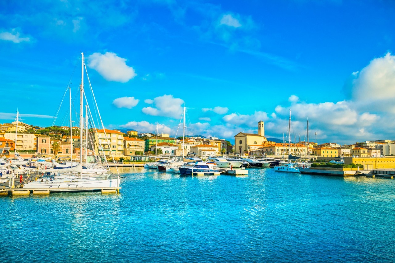 View of a marina. Several boats can be seen in the bay on the water and the coastal town with its residential houses can be seen in the background 