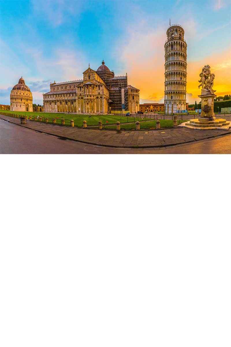 View from a bend in the road across a lawn to three white monumental buildings in the evening. On the left of the picture is a round baptistery, to the right of it, in the centre, a cathedral and next to that a leaning tower 
