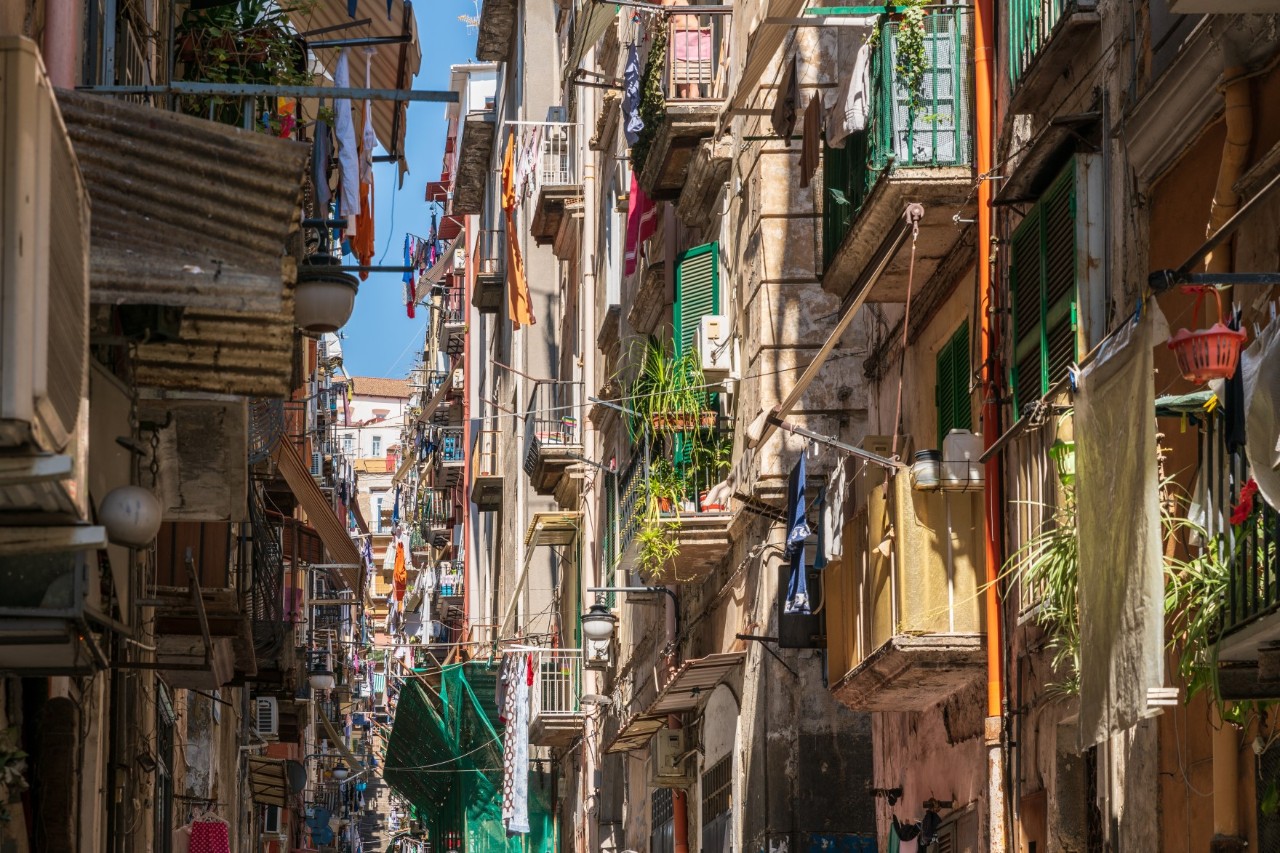 A narrow street in Naples’ Old Town with old buildings, planted balconies and washing lines right and left. The sky is blue.