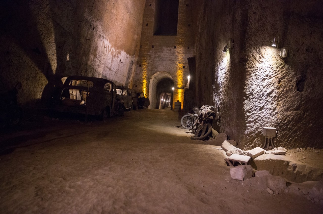 An underground tunnel illuminated by several lamps. On the left are old, dilapidated cars and a scrapped motorbike on the right. 