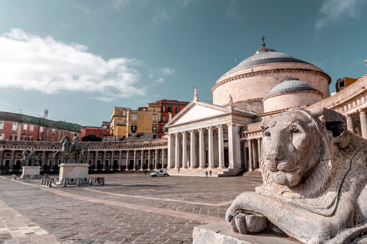 Quiet square with church in the background, tympanum and pillared arch in front. On the right is a stone lion’s head on a pedestal.