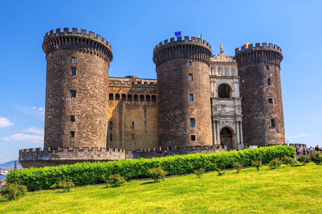 A fortress with three massive towers. There is a green hedge and lawn in the foreground and blue sky on the horizon. 