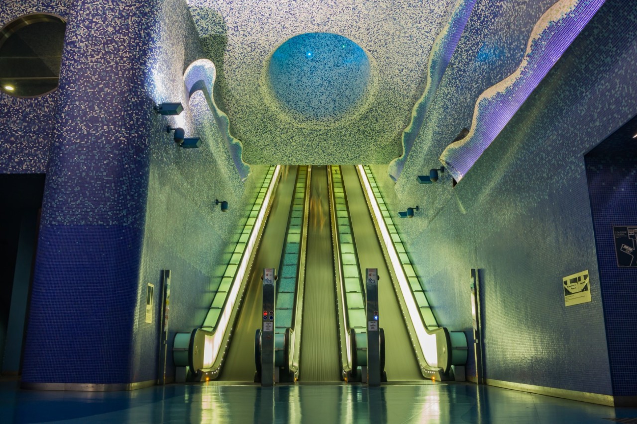 A worm’s-eye view of an underground station. Two escalators are illuminated in green and there are round and wave-shaped light installations in shades of blue on the ceiling and walls. © Bruno Coelho/stock.adobe.com