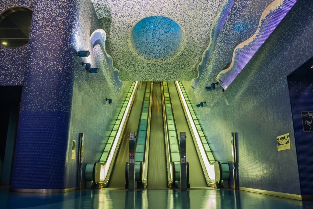 An underground station. Two escalators are illuminated in green and there are round and wave-shaped light installations in shades of blue on the ceiling and walls. 