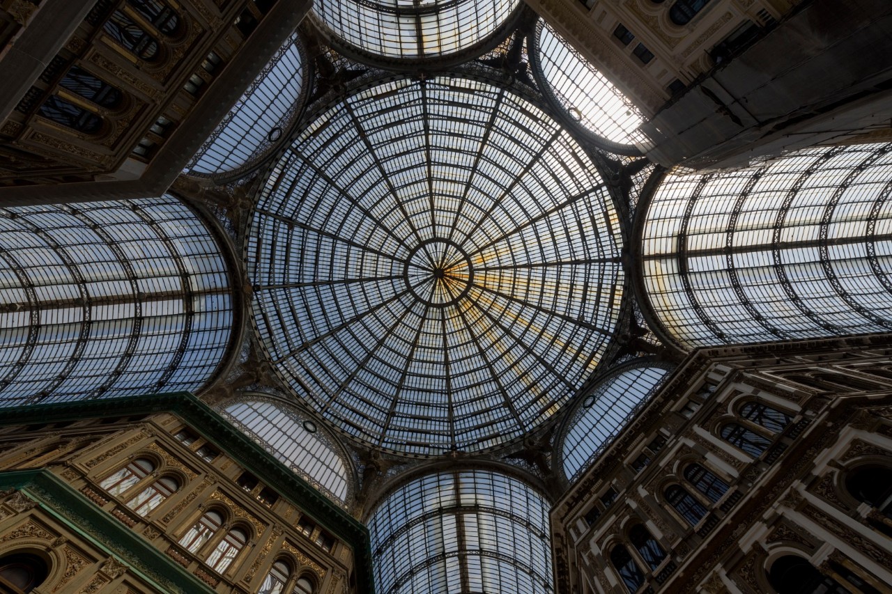 A shopping arcade ceiling with a glass dome resembling a wheel, with glass columns leading down from the dome.