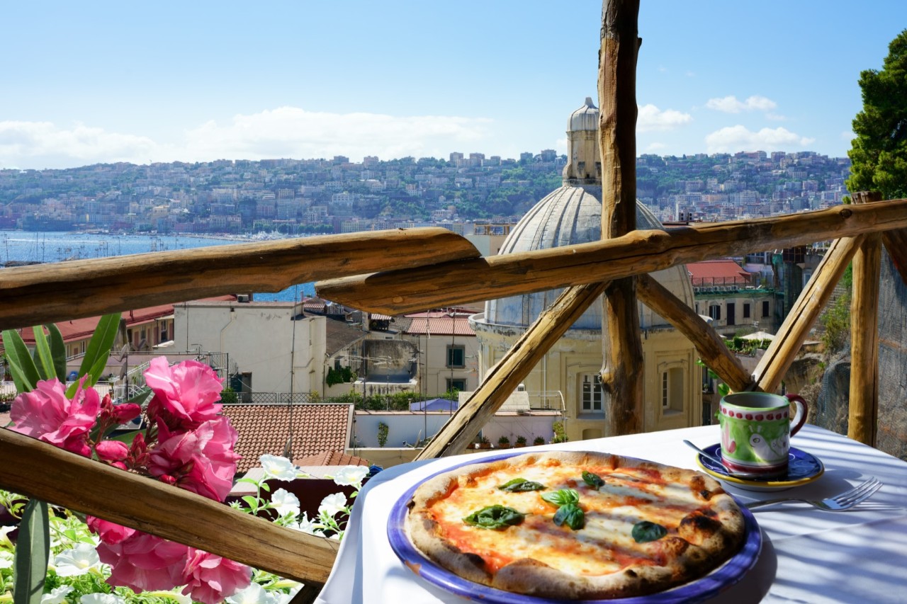 A restaurant terrace overlooking the rooftops of Naples with a table laid with a white cloth, pizza and coffee cup. In the background, a church dome, buildings and the sea to the left.