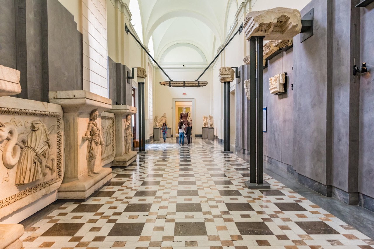 An archaeological museum. On the left are reliefs on the wall, at the end of the corridor various sculptures and visitors. The floor is a tiled mosaic pattern.