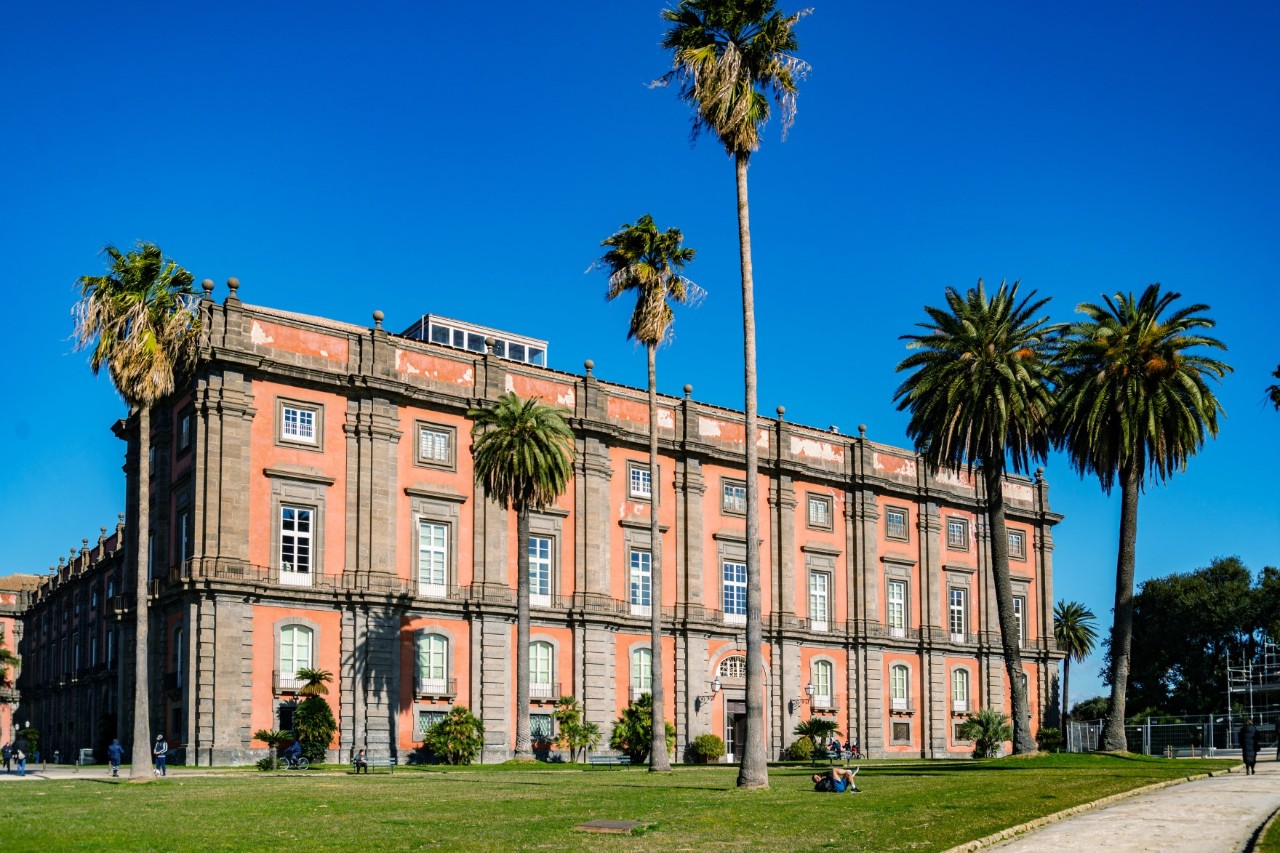 Three-storey, bright red building in neoclassical style lined with palm trees, bright blue sky in the background. 