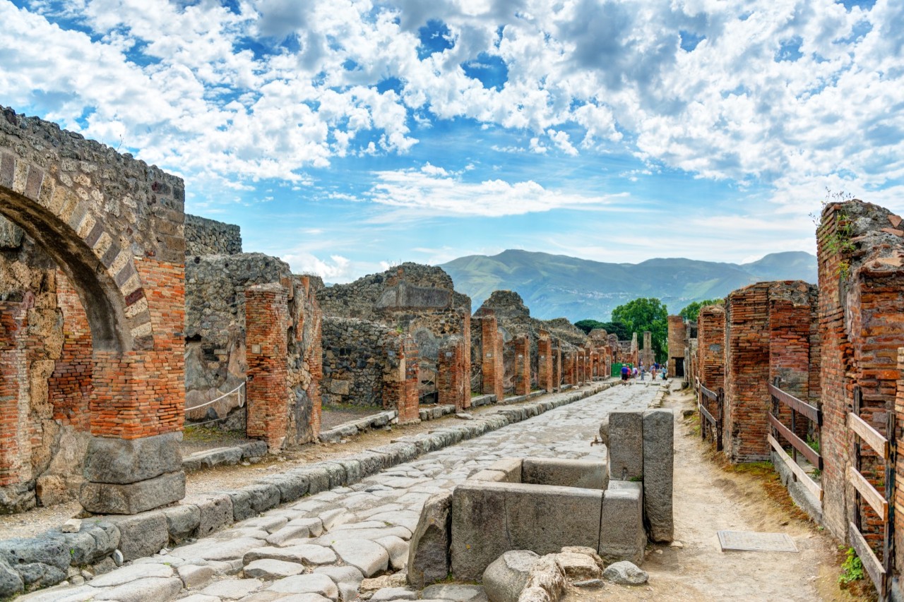 An ancient street in Pompeii, lined with ruins. Volcanic mountains and skies with cumulus clouds can be seen on the horizon.  