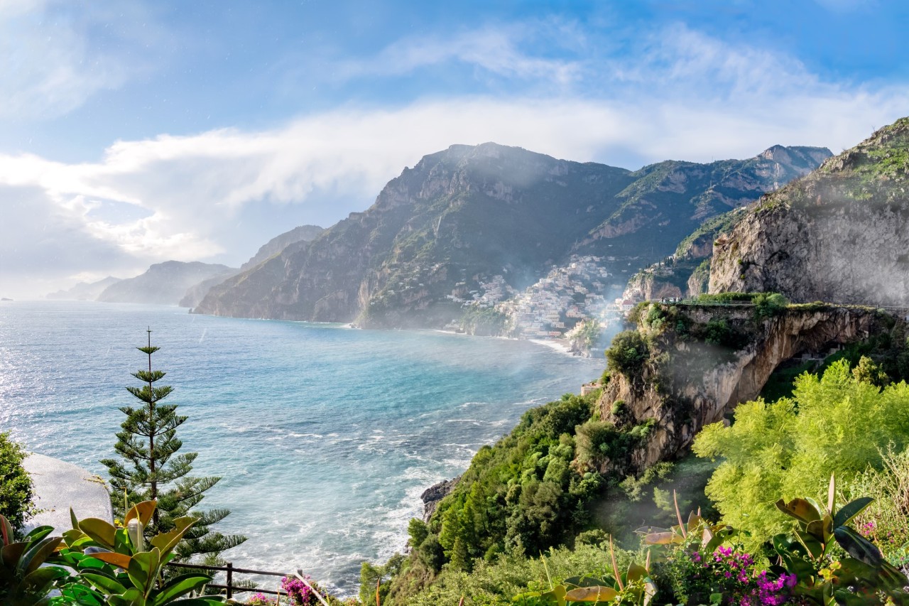 View of a stretch of the Amalfi Coast. Blue sea meets rocky coastline, the blue sky is lightly streaked with clouds. In the foreground are trees and flowers. © refresh(PIX)/stock.adobe.com