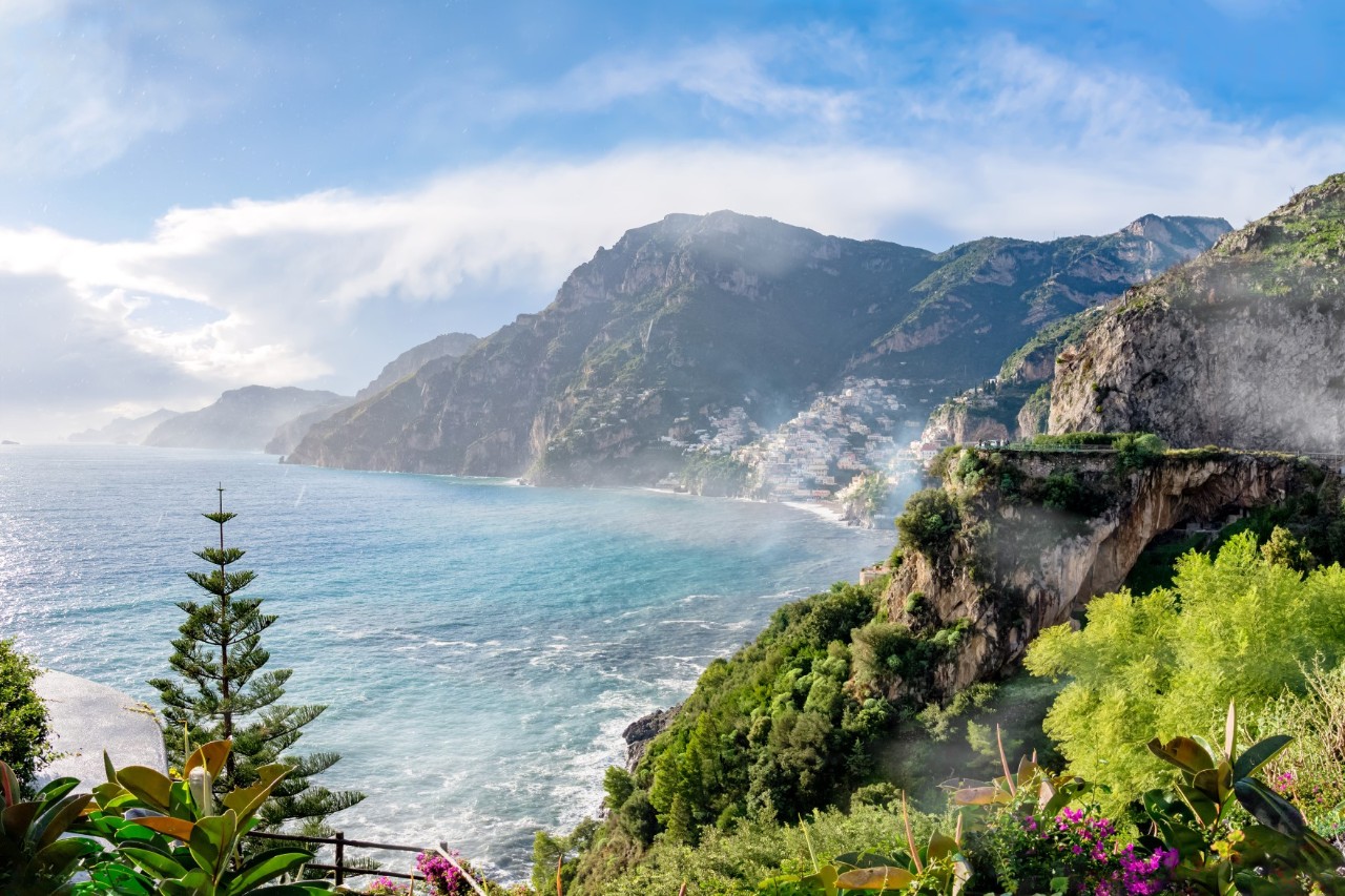 A stretch of the Amalfi Coast. Blue sea meets rocky coastline, the blue sky is lightly streaked with clouds. In the foreground are trees and flowers.