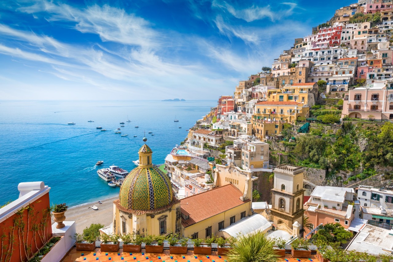 View of colourful houses on a mountain slope, church dome in the foreground, terrace with flower boxes, turquoise bay with boats on the left.© IgorZh/stock.adobe.com