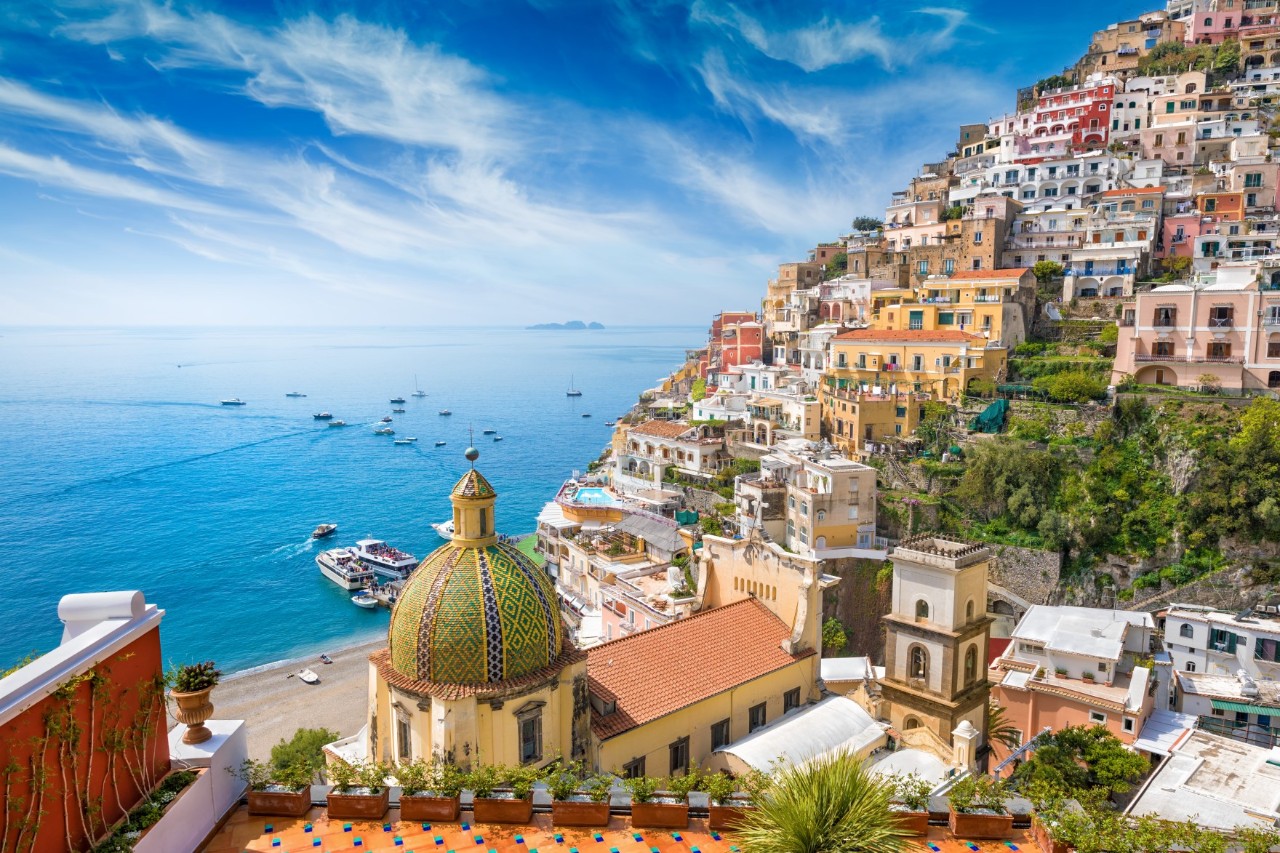 Colourful houses on a mountain slope, church dome in the foreground, terrace with flower boxes, turquoise bay with boats on the left.