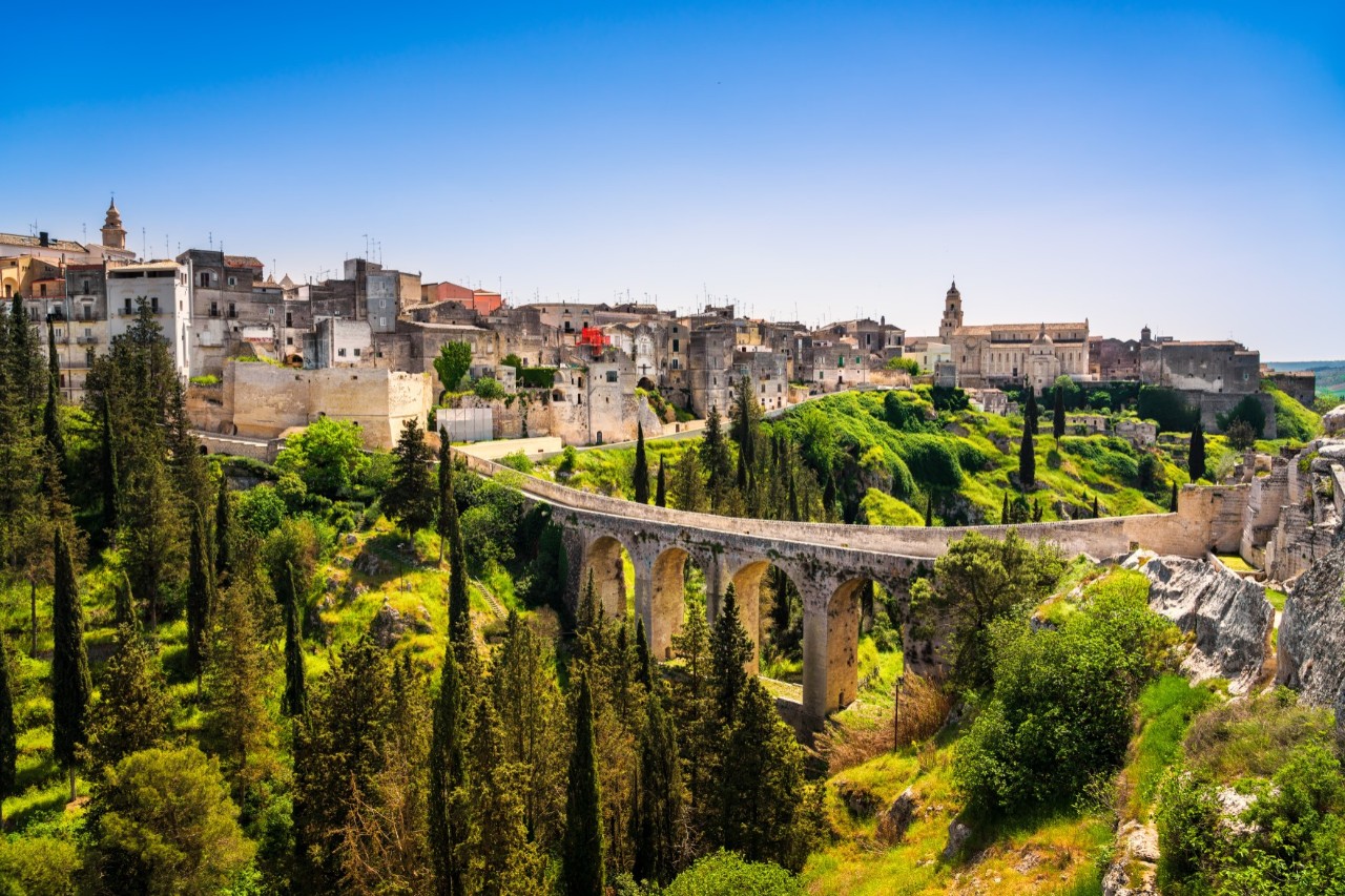 A mighty stone bridge with round arches spans a valley, behind which lies a small Italian town.
