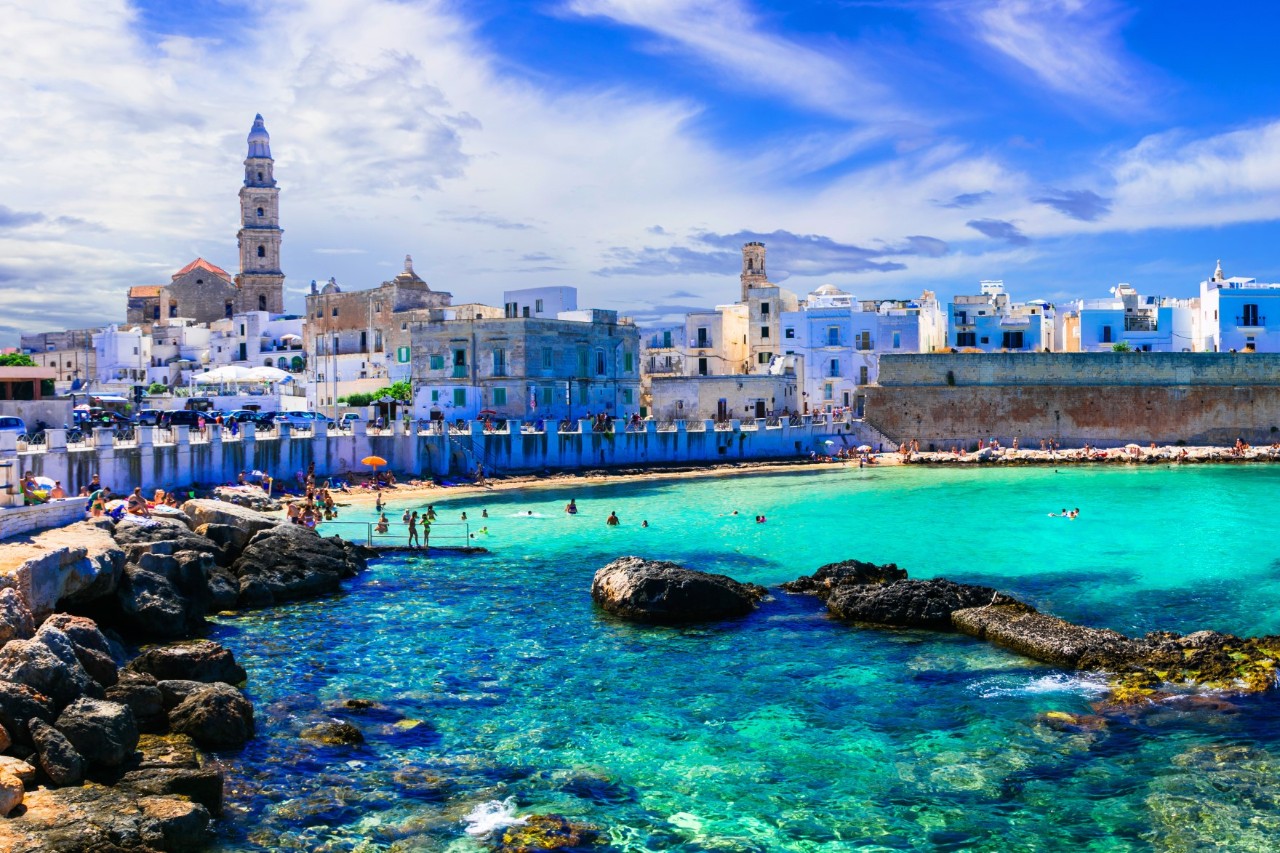 Bay with turquoise blue water and rocky shore. In the background is a Mediterranean town with white buildings and a city wall.